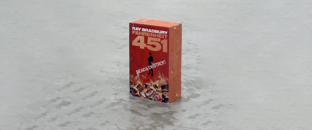 Das Bild zeigt ein Kunstwerk des Kollektivs Claire Fontaine: Ein Backstein umhüllt vom Cover des Buches Fahrenheit 451 von Ray Bradbury.