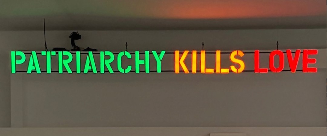 Das Bild zeigt ein Kunstwerk des Kollektivs Claire Fontaine: Ein großer Neonschriftzug mit den Worten "Patriarchy kills love" hängt in einem großen Ausstellungsraum oberhalb einer Art weißen Tribüne.